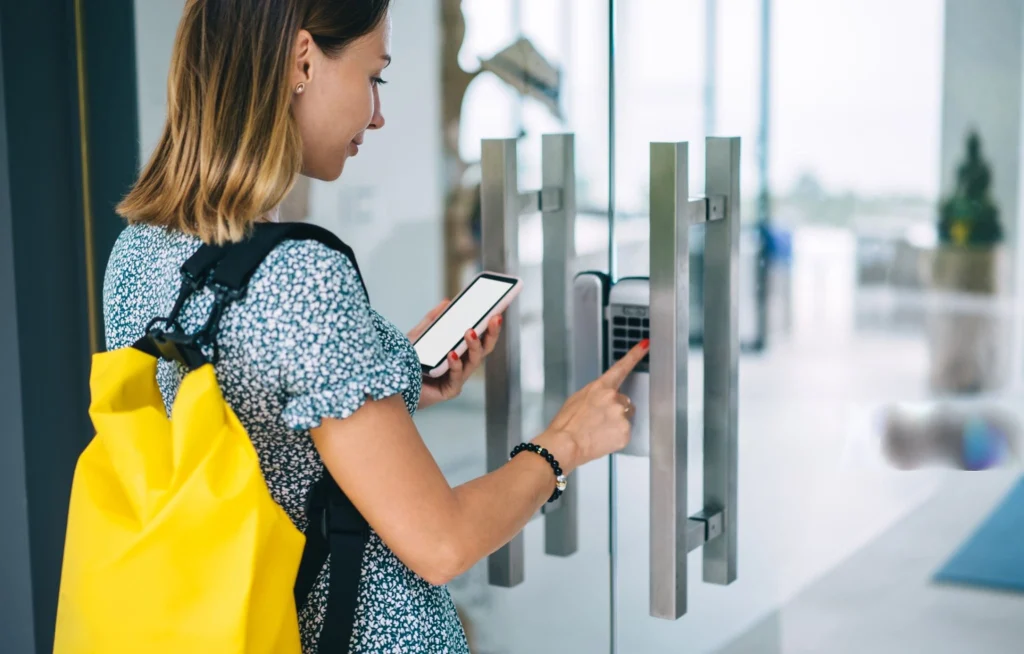 Women using a keypad access control system with a smartphone in hand at a glass door.